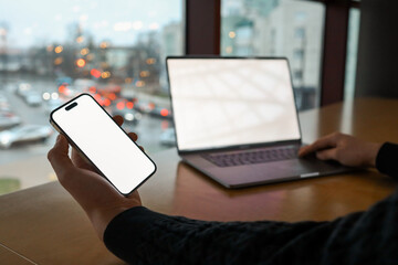 Person holding a smartphone with a blank white screen in front of an open laptop at a wooden desk near a window. Modern workspace, technology, and digital connectivity concept