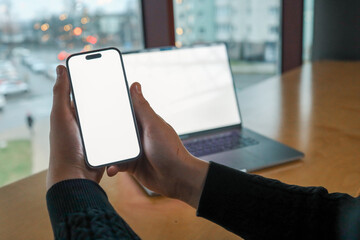 Person holding a smartphone with a blank white screen in front of an open laptop at a wooden desk near a window. Modern workspace, technology, and digital connectivity concept