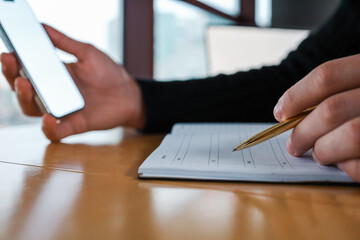 Person holding a smartphone and writing in a planner with a gold pen at a wooden table near a window. Modern workspace, multitasking, and digital planning concept in a professional setting