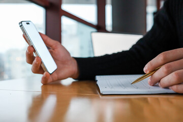 Person holding a smartphone and writing in a planner with a gold pen at a wooden table near a window. Modern workspace, multitasking, and digital planning concept in a professional setting