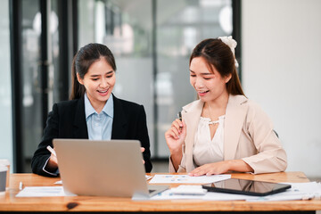 Fototapeta premium Two businesswomen working together in an office, discussing documents and using a laptop, showcasing teamwork and collaboration.