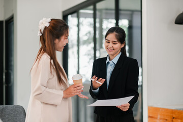 Two professional women discussing business in a modern office, holding documents and coffee, showcasing teamwork and collaboration.