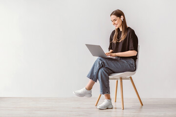 Cheery young woman sitting on chair, using laptop computer, wearing earphones, studying remotely or having video call against white studio wall, blank space. Millennial lady watching movie online