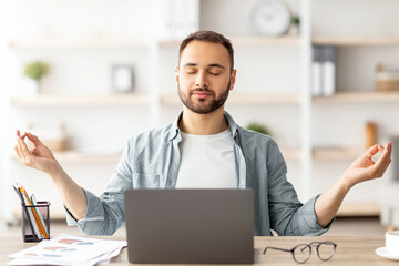 Workplace stress management. Calm Caucasian man meditating with closed eyes in front of laptop pc...