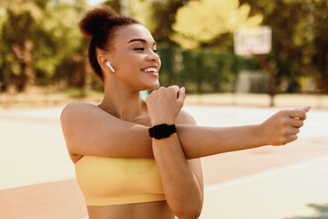 Warm Up Concept. Portrait of cheerful young slim black woman stretching her arms while standing outdoors at public playground, smiling fit lady wearing bright yellow sportswear, earbuds and smartwatch