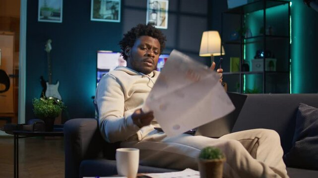 Black freelancer does his research by reading articles and data driven papers at home, using phone and pc to collect information for a new project. Man multitasking and studying files. Camera B.