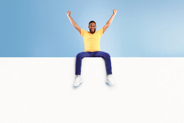 Fototapeta premium Advertising billboard template. Young African American guy sitting on top of blank white poster with mockup, lifting hands up in excitement on blue studio background, space for design