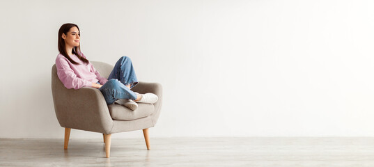 Young Caucasian woman relaxing in cozy armchair against white studio wall, copy space. Peaceful...