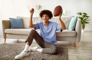 Excited black teenager watching American football on television at home, full length. Emotional teen guy cheering for his favorite rugby team, following international championship on TV