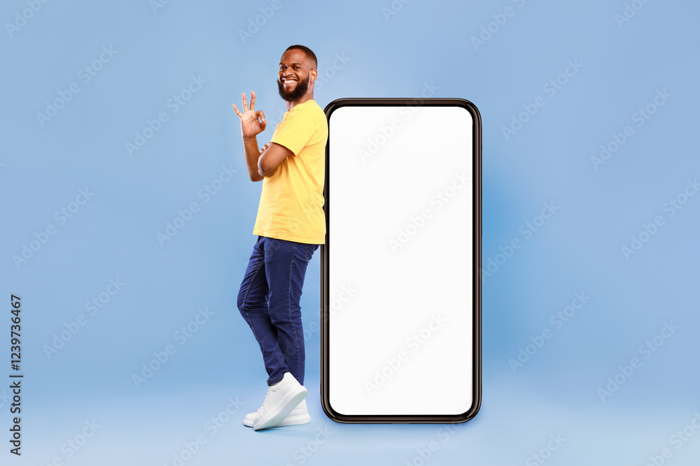 Poster optimistic african american man showing ok gesture, leaning on huge phone with blank white screen, r