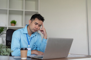 Pensive Asian businessman deep in thought at his desk with a laptop and coffee cup in a modern office environment.
