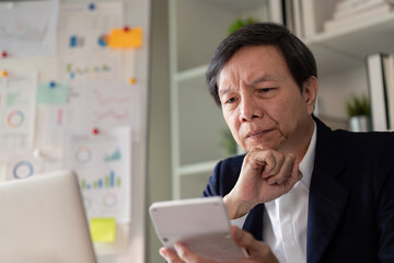 Fototapeta premium Elderly Asian businessman analyzing data on a smartphone while working in a modern office environment.