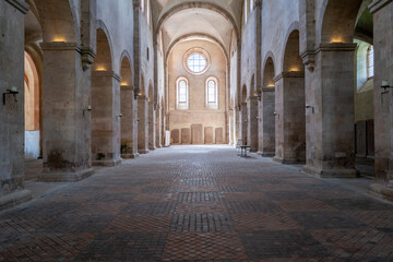 Sunlight streams into a serene ancient abbey, illuminating the stone columns and brick floor.  A peaceful, historical scene.