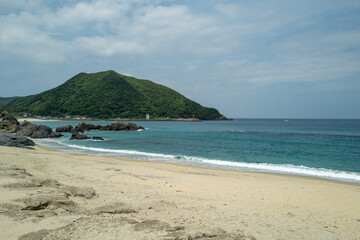 A tranquil beach scene: clear turquoise water gently rolling onto a sandy shore, with a verdant hill rising in the background.