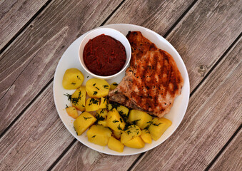 A plate of delicious lunch, grilled pork entrecote, boiled potatoes with dill and a cup of sauce on a wooden table.