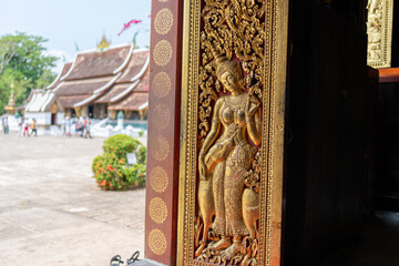 Intricate golden carving of a woman graces a temple door in Laos, the courtyard visible beyond.