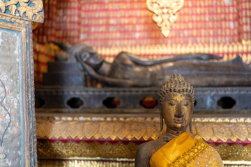 Golden Buddha statue sits serenely before a reclining Buddha in a beautifully ornate, red-gold Thai...