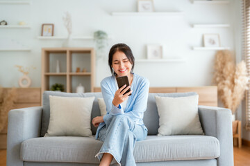 Smiling pretty young Asian woman sitting on cozy couch, using funny mobile apps in living room. Woman at Home, Doing Online Shopping, Messaging Friends, Posting on Social Media