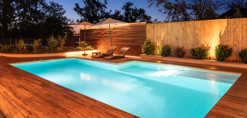 Illuminated backyard swimming pool with wooden deck and lounge chairs. Serene outdoor space surrounded by greenery and soft lighting