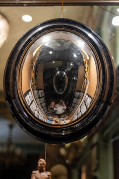 Two people take a selfie reflected in a unique convex mirror surrounded by framed artwork in a shop.