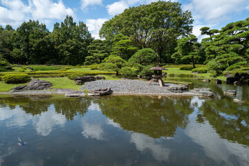 Fototapeta premium A tranquil Japanese garden scene with a stone lantern beside a peaceful pond, surrounded by lush greenery under a blue sky.