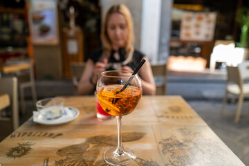 A refreshing Aperol Spritz sits on a cafe table, bathed in sunlight; a woman sits blurred in the...
