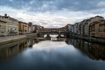 Obraz premium The iconic Ponte Vecchio in Florence, Italy, gracefully spans the Arno River, reflecting the city's charm under a tranquil sky.
