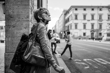 Naklejka premium A thoughtful senior woman pauses on a busy city street, looking up at the buildings, carrying multiple bags.