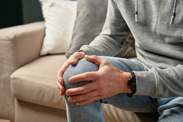 Close-up of man gripping his knee in discomfort while sitting on couch. Man in casual clothing...