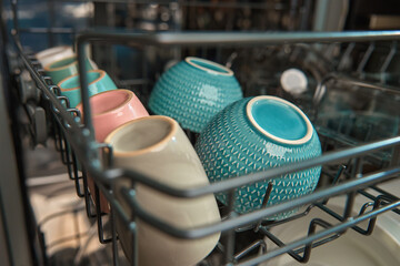 Close-up view of colorful ceramic bowls and cups neatly arranged inside dishwasher rack. Modern home appliance ensuring efficient dish cleaning