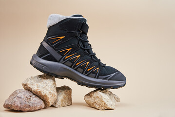 Black hiking boot with orange details on small rocks. Close up of waterproof trekking shoe on beige...