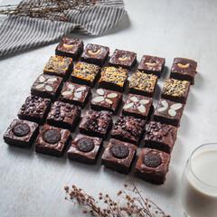 Assorted brownies with different toppings are neatly arranged on a surface. A glass of milk sits nearby, adding to the indulgent appeal