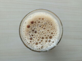 A top-down view of a glass of coffee with a frothy and bubbly surface, placed on a light-colored table.