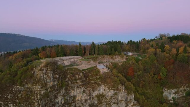 A breathtaking aerial shot capturing the historic Punta Corbin Fort nestled within vibrant autumn forested hills, with a serene pink sky providing a stunning backdrop.