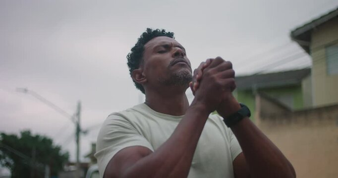 Man of African descent praying intensely, hands clasped, head tilted upward, deep spiritual devotion, seeking strength and guidance, urban background, moment of faith and resilience