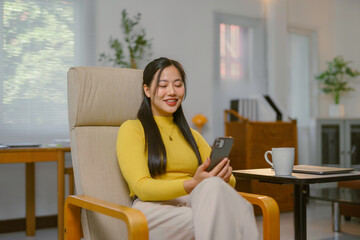 Obraz premium Young asian woman using smartphone sitting on armchair at home