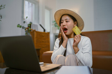 Surprised asian woman holding credit card and using laptop for shopping online at home