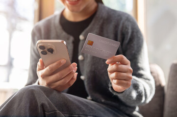 A close-up of a woman sits indoors on a bright day, holding a smartphone and a credit card.