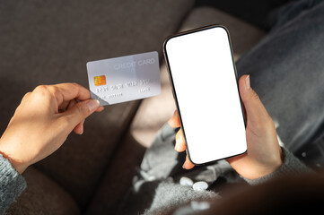 Top view of a woman sits indoors, holding a credit card and a smartphone with a white screen mockup.