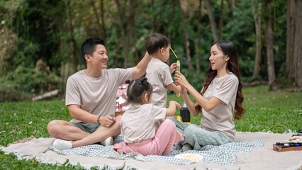 A lovely Asian family is enjoying a picnic in a park, with the mom and dad playfully blowing bubbles