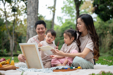 A happy Asian family enjoys painting on a canvas during a picnic in a public park.