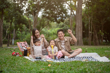 A happy, cheerful Asian family sits on a picnic blanket in a public park, smiling at the camera.