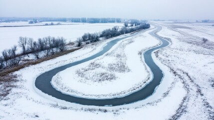 Fototapeta premium Snowfall aerial capture of a frozen river winding through a snowy landscape