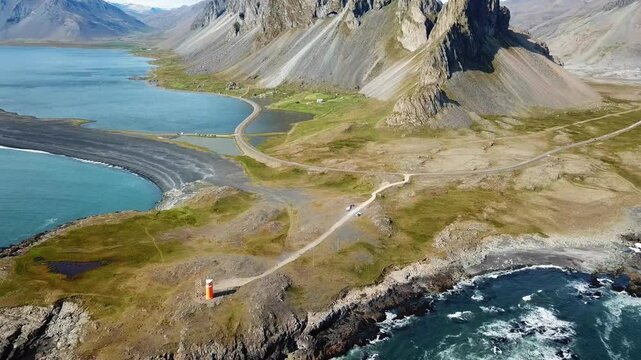 Breathtaking aerial view of Hvalnes Nature Reserve in Iceland, featuring a winding road to an orange lighthouse, surrounded by rugged mountains and a tranquil lagoon, drone establishing shot