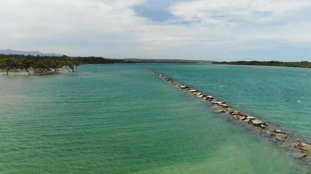 Bellinger River with rock barrier dividing calm turquoise waters, surrounded by natural greenery, perfect for travel and nature visuals, Urunga, New South Wales, Australia. Aerial forward