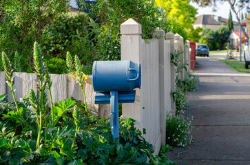 A mailbox against a traditional white picket fence, surrounded by lush garden plants along a suburban sidewalk. A quintessential Australian residential neighborhood in Melbourne, Australia.