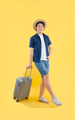 Full body image of a young Asian man pulling a suitcase, wearing a travel camera and posing on a yellow background