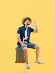 Full body image of a young Asian man sitting on  a suitcase, wearing a travel camera and posing on...