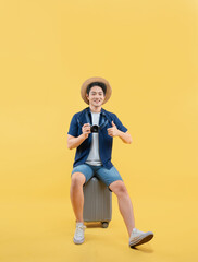 Full body image of a young Asian man sitting on  a suitcase, wearing a travel camera and posing on a yellow background