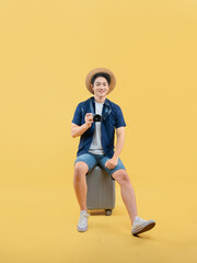 Full body image of a young Asian man sitting on  a suitcase, wearing a travel camera and posing on a yellow background